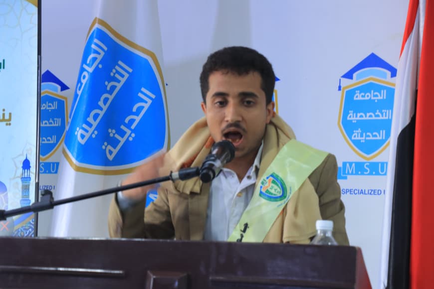 IMG-20260907-WA0039 - جامعة 21 سبتمبر Man in a beige suit speaks into a microphone at a podium, with blue banners and flags behind him at a conference.