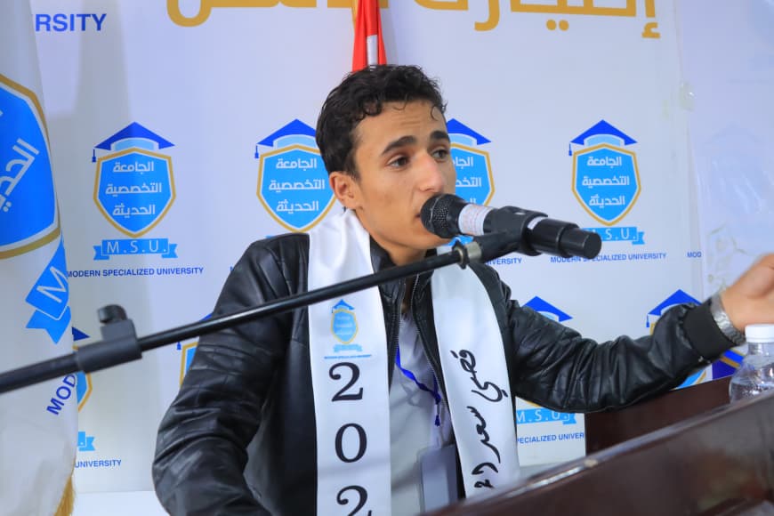 IMG-20260907-WA0041 - جامعة 21 سبتمبر Student speaker at a university event, wearing a white sash marked 2022 and speaking into a microphone at a podium, with MSU logos in the background.