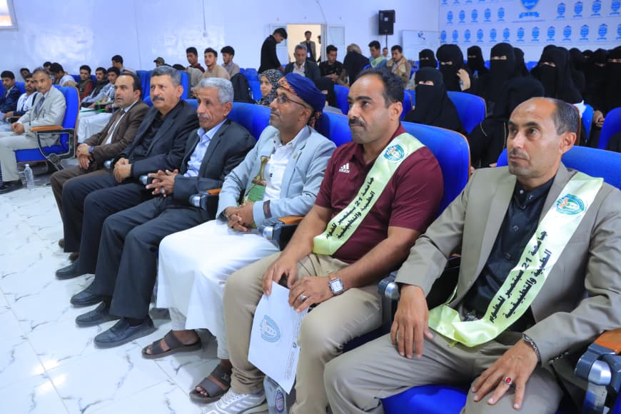 IMG-20260907-WA0043 - جامعة 21 سبتمبر Front row of attendees at a formal event in blue seats, several men in suits and traditional dress wearing light green sashes with writing.