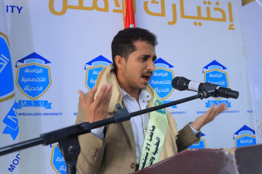 IMG-20260907-WA0045 - جامعة 21 سبتمبر Young man speaking at a podium during a university event, gesturing with his hands, with blue and gold university banners in the background.