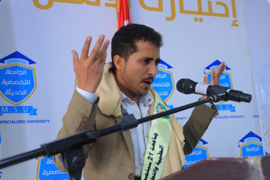 IMG-20260907-WA0047 - جامعة 21 سبتمبر Man in a beige blazer and scarf speaks at a podium, raising both hands in a speech, with a university banner in Arabic behind him.