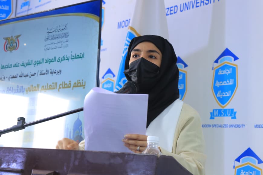 IMG-20260907-WA0048 - جامعة 21 سبتمبر Speaker wearing a hijab and face mask delivering a speech at a podium, holding papers with a university banner in the background.