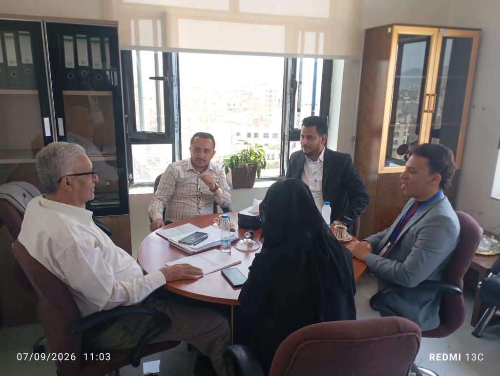 Five professionals sit around a round conference table in a bright office, reviewing documents and devices during a meeting.