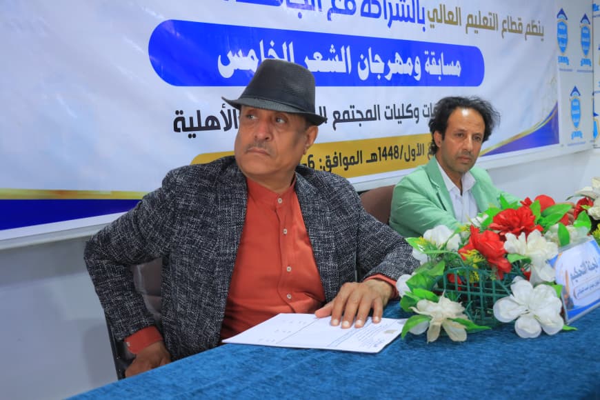 IMG-20260907-WA0049 - جامعة 21 سبتمبر Two men seated at a table during a formal event; the man in the foreground wears an orange shirt and gray fedora, with a blue banner and a flower arrangement in front of them.