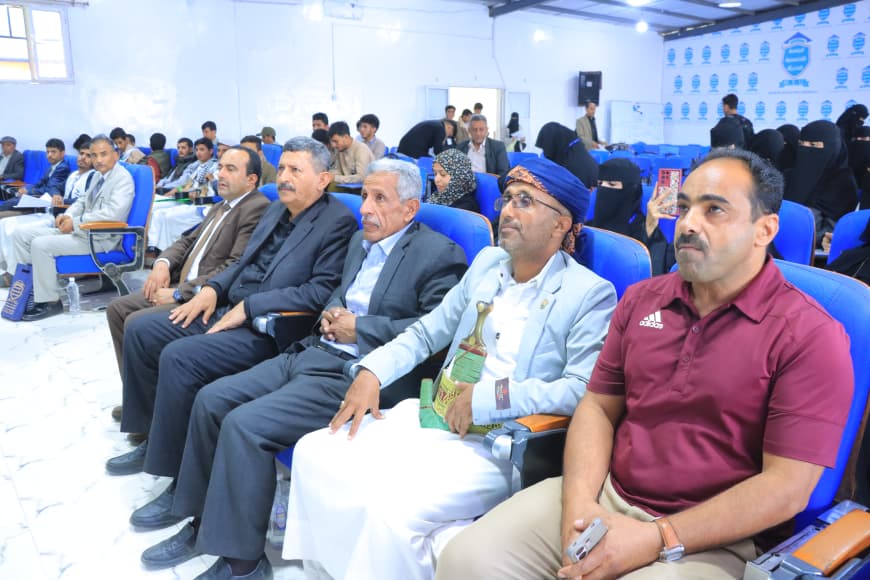 IMG-20260907-WA0050 - جامعة 21 سبتمبر Group of men in suits and traditional clothing seated in blue conference chairs, listening to a presentation in a well-lit hall with attendees in the background.