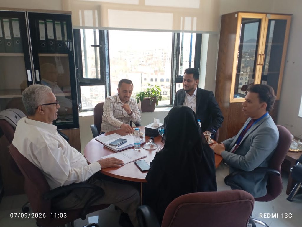 Five professionals in a bright office around a round table, reviewing documents and discussing; a woman wearing a hijab faces away as others listen.