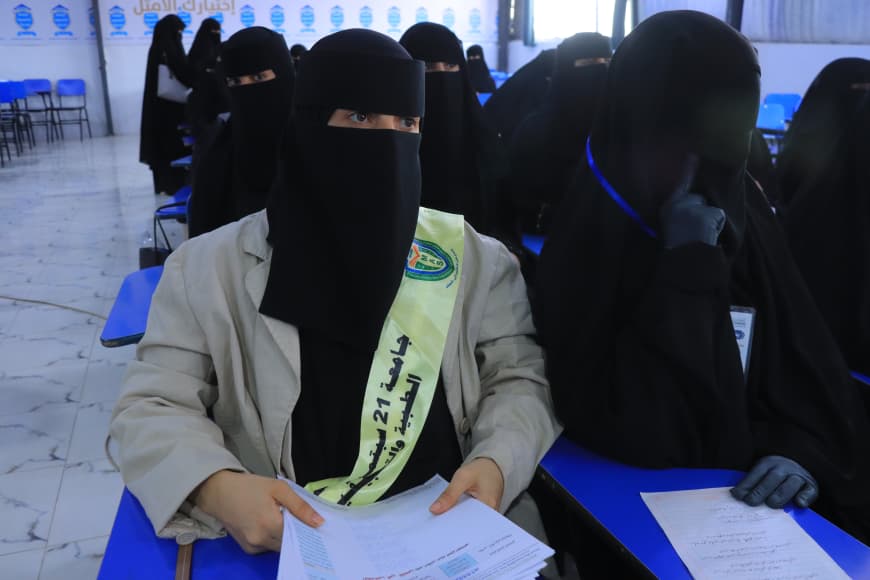 IMG-20260907-WA0051 - جامعة 21 سبتمبر Woman in a beige blazer and niqab with a pale sash seated at a blue desk, holding papers in a classroom setting.