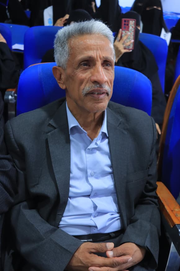 IMG-20260907-WA0052 - جامعة 21 سبتمبر Older man with gray hair and a mustache in a dark suit, seated in a blue chair at an event area.