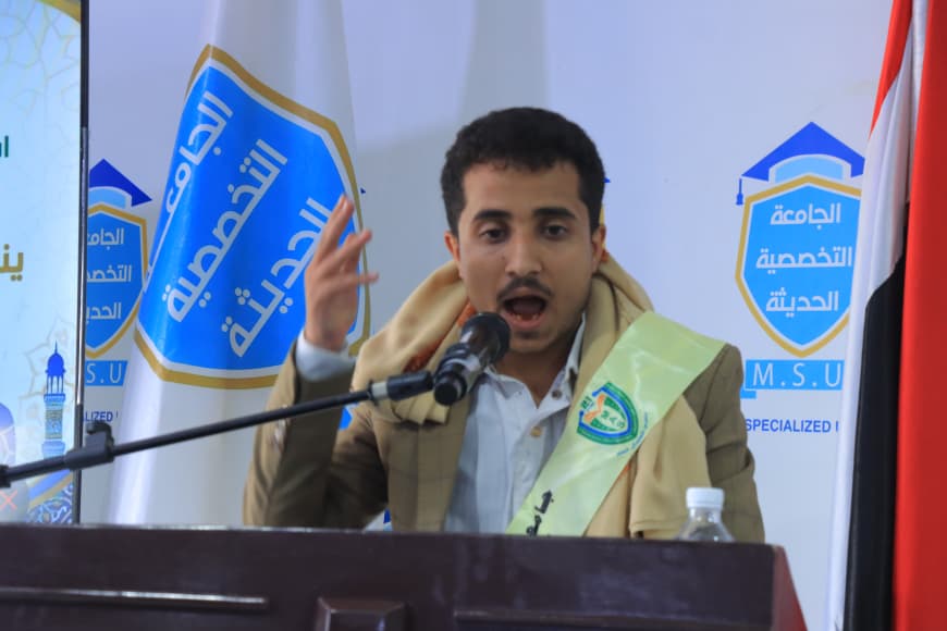 IMG-20260907-WA0053 - جامعة 21 سبتمبر Man speaking at a podium, wearing a beige scarf, with a microphone and banners with Arabic text in the background.