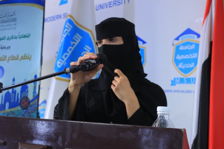 IMG-20260907-WA0055 - جامعة 21 سبتمبر Woman in a black niqab speaks into a microphone at a podium during a formal event, with blue university logos in the background.”