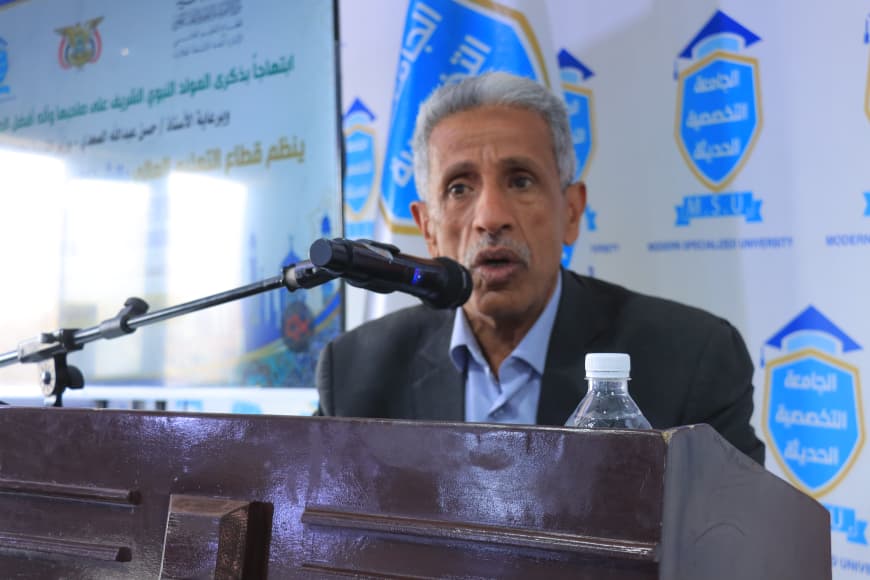 IMG-20260907-WA0056 - جامعة 21 سبتمبر A man in a suit speaks into a microphone at a wooden podium, with a water bottle on the lectern and blue banners in the background.
