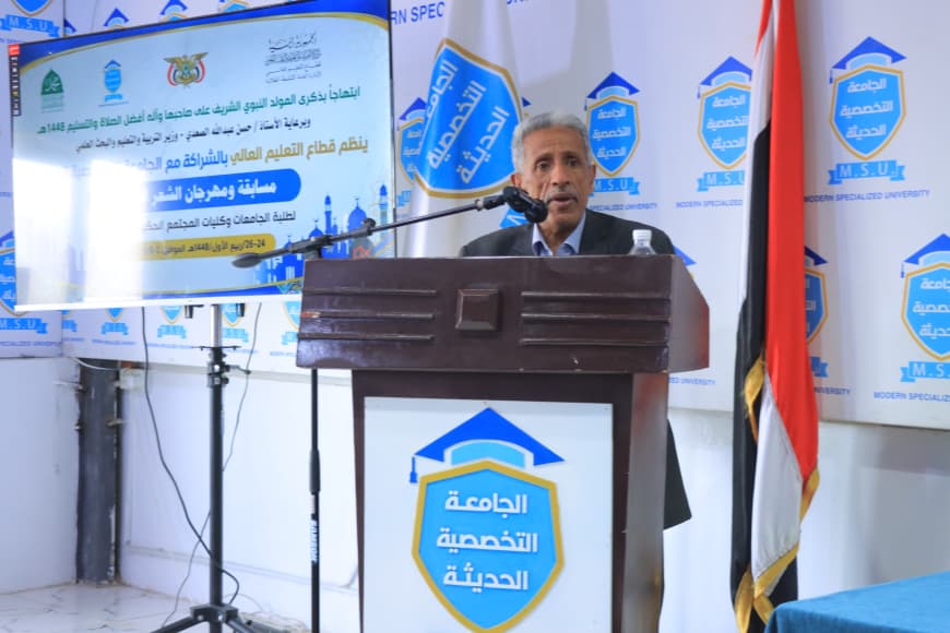 IMG-20260907-WA0057 - جامعة 21 سبتمبر Man speaks at a podium during a formal university event, with blue banners and an Arabic shield logo in the background and an Egyptian flag nearby.