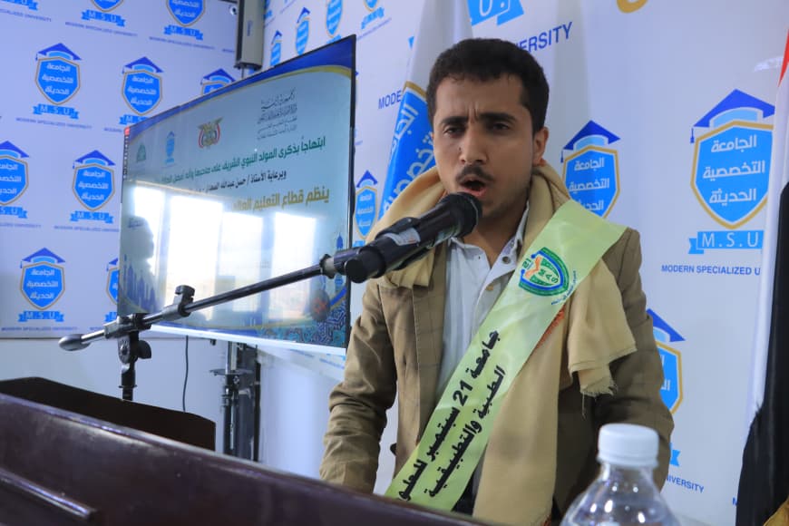 IMG-20260907-WA0058 - جامعة 21 سبتمبر Man speaking at a podium with a microphone during a formal event, wearing a scarf and sash, with blue branded banners in Arabic in the background.