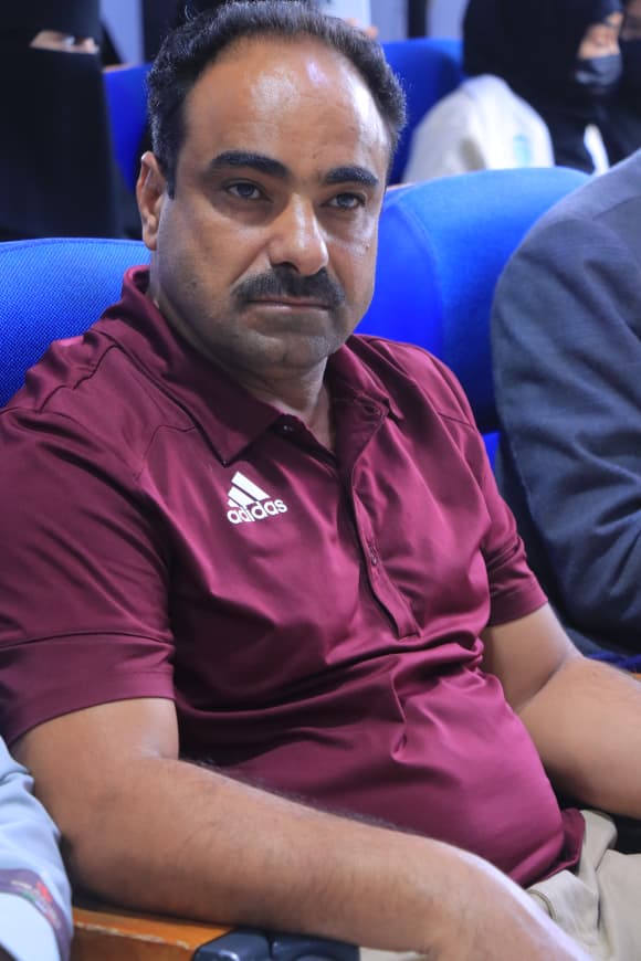 IMG-20260907-WA0062 - جامعة 21 سبتمبر Man in a maroon Adidas polo sitting in blue auditorium seats, looking toward the camera at an event