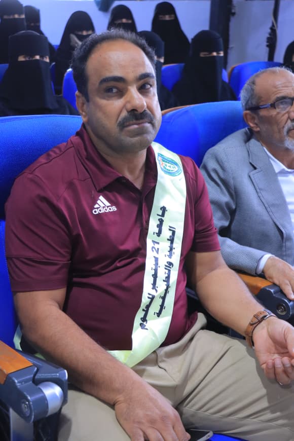 IMG-20260907-WA0063 - جامعة 21 سبتمبر Man in a maroon polo seated in blue auditorium chairs, wearing a light sash with Arabic text at an event, with people in background.