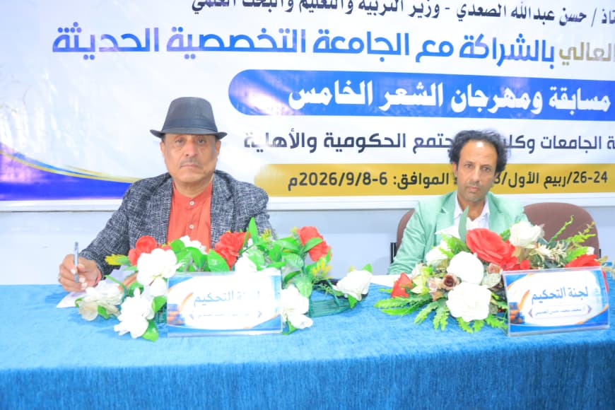 IMG-20260907-WA0065 - جامعة 21 سبتمبر Two men seated at a blue table with flower arrangements during a formal event, with a large Arabic banner behind them referencing a university specialization festival.