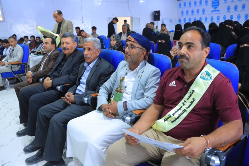 IMG-20260907-WA0067 - جامعة 21 سبتمبر Group of men in suits seated in blue auditorium chairs at a formal ceremony; a man in maroon shirt with a sash sits front right, while women in black niqab are in the background near a blue-patterned wall.