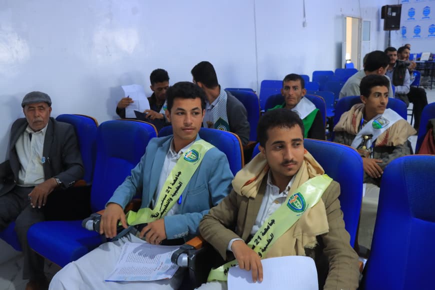 IMG-20260907-WA0068 - جامعة 21 سبتمبر Two young men in light blazers wearing sashes sit in blue auditorium chairs, holding papers and a microphone, at an event.