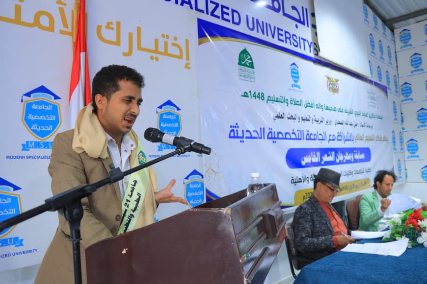 IMG-20260907-WA0069 - جامعة 21 سبتمبر Man in a beige coat speaks at a wooden podium with a microphone during a university event, with a large Arabic banner and two panelists seated beside him.
