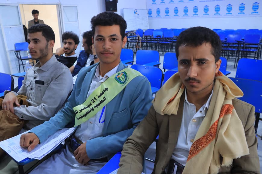 IMG-20260907-WA0070 - جامعة 21 سبتمبر Four young men sit in a classroom with blue chairs; the central one wears a light-blue jacket and sash, looking at the camera, with two others nearby and more attendees in the background.