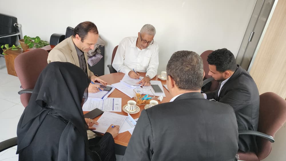 IMG-20260908-WA0033 - جامعة 21 سبتمبر Five professionals around a wooden table reviewing documents and taking notes in a bright office setting, with cups and tablets nearby (team meeting).