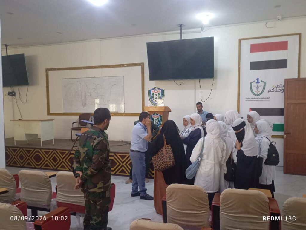 Group of students in white hijabs gathered near a stage, with a speaker and a soldier nearby in a classroom auditorium.