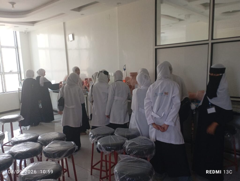 Group of women in white lab coats and hijabs gather at a clinic counter for a training session in a clinical skills lab.