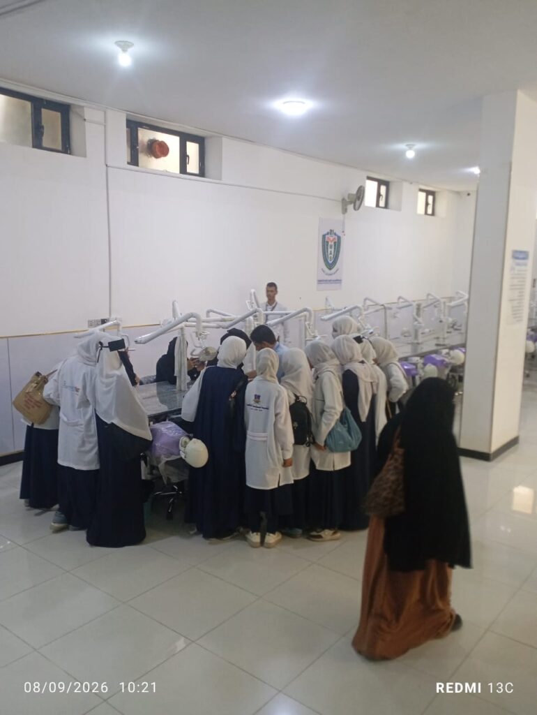 Group of female students in white coats and hijabs gathered around a row of dental chairs in a clinic, with an instructor at the front.