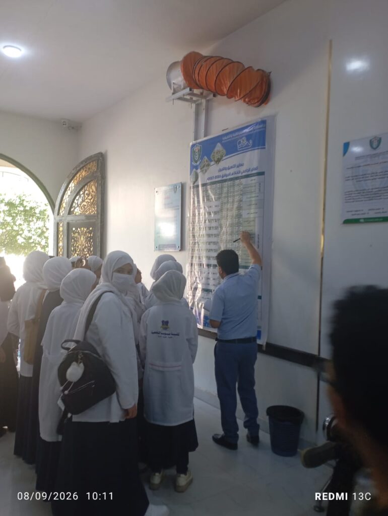 Group of female students in white coats and hijabs gathered around a large poster on the wall as a man points to it for explanation in a hallway.