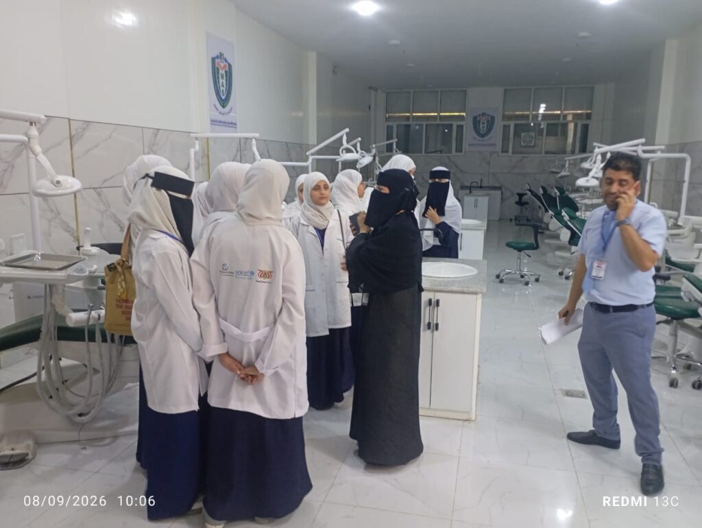 Group of women in white coats and hijabs listening to a woman in black outside a dental chair in a bright clinic.