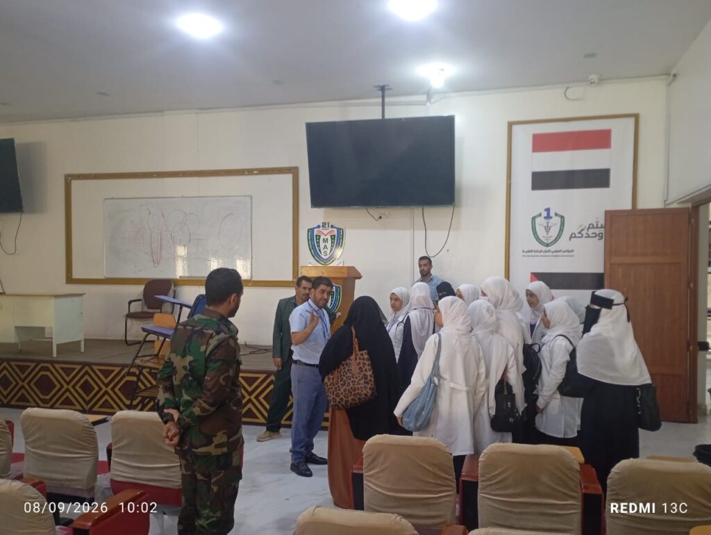 Group of young women in white hijabs cluster around a speaker in a classroom-style hall; a soldier stands nearby.