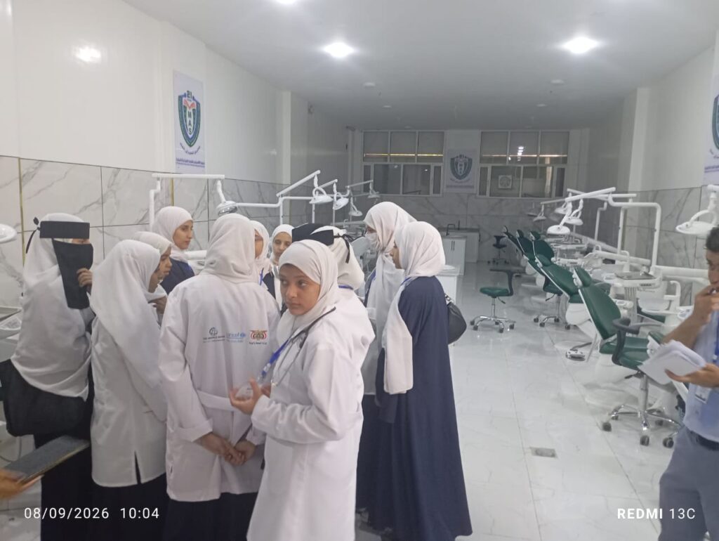 Group of female students in white coats and hijabs gathered in a bright dental clinic, listening to a instructor near dental chairs and equipment.