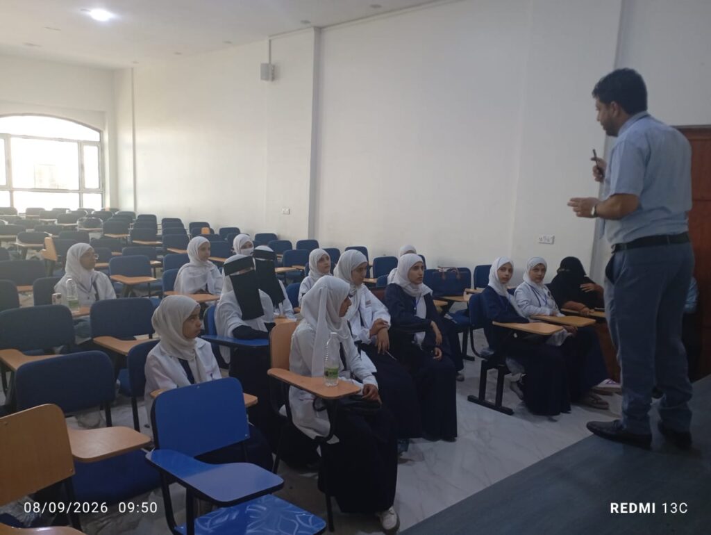 Male instructor lecturing at the front of a classroom to rows of female students wearing hijabs seated with desks.