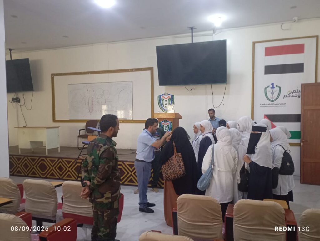 Group of women in white hijabs lined up to speak with a presenter at a podium in a classroom-style hall, with a soldier in camouflage nearby.