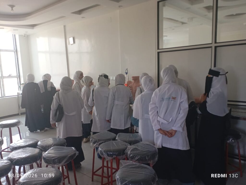 Group of women in white lab coats and hijabs gathered in a clinic, listening at a counter near a glass partition.