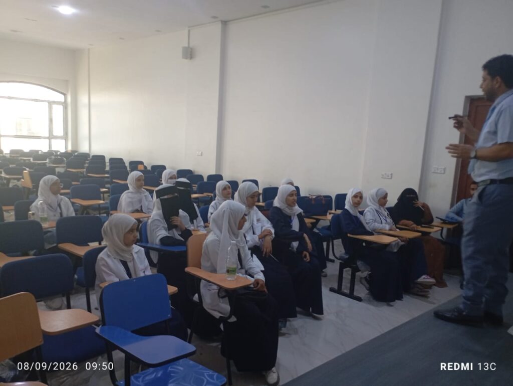 Instructor at the front lecturing to female students wearing hijabs in a bright classroom filled with blue and orange desks.