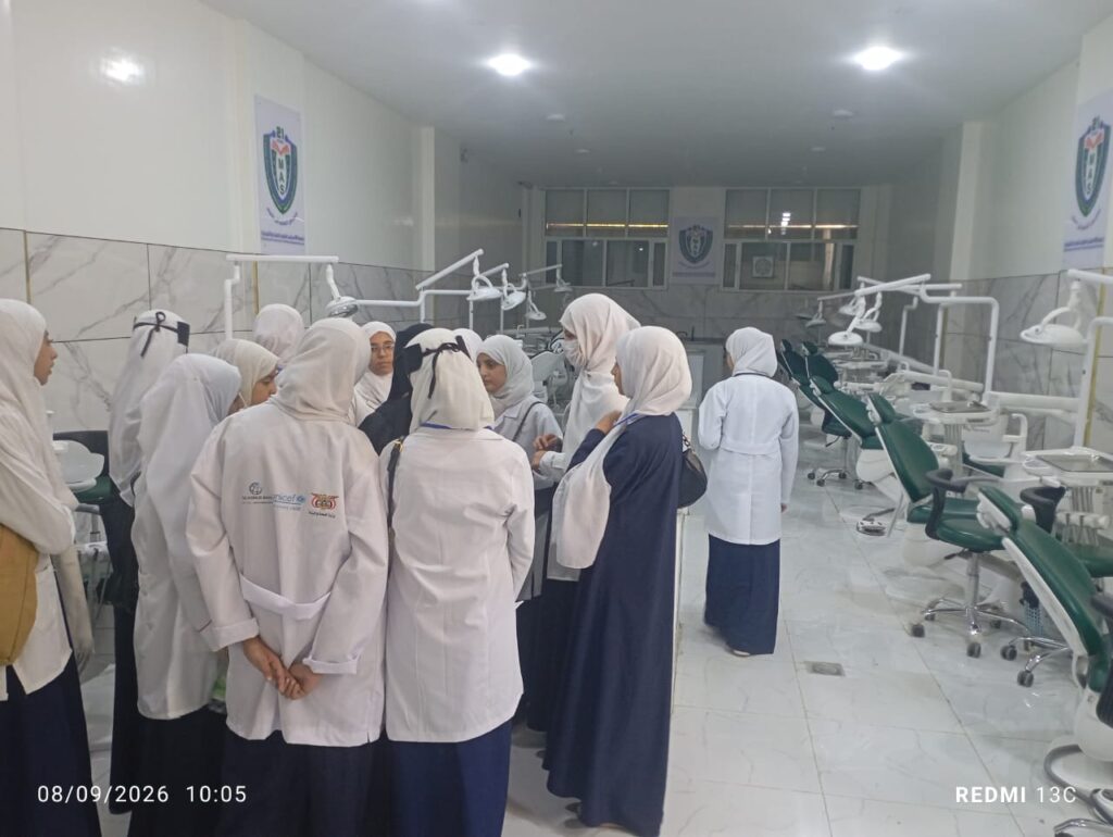 Group of people in white lab coats and hijabs gathered in a dental clinic, discussing near row of dental chairs and equipment.