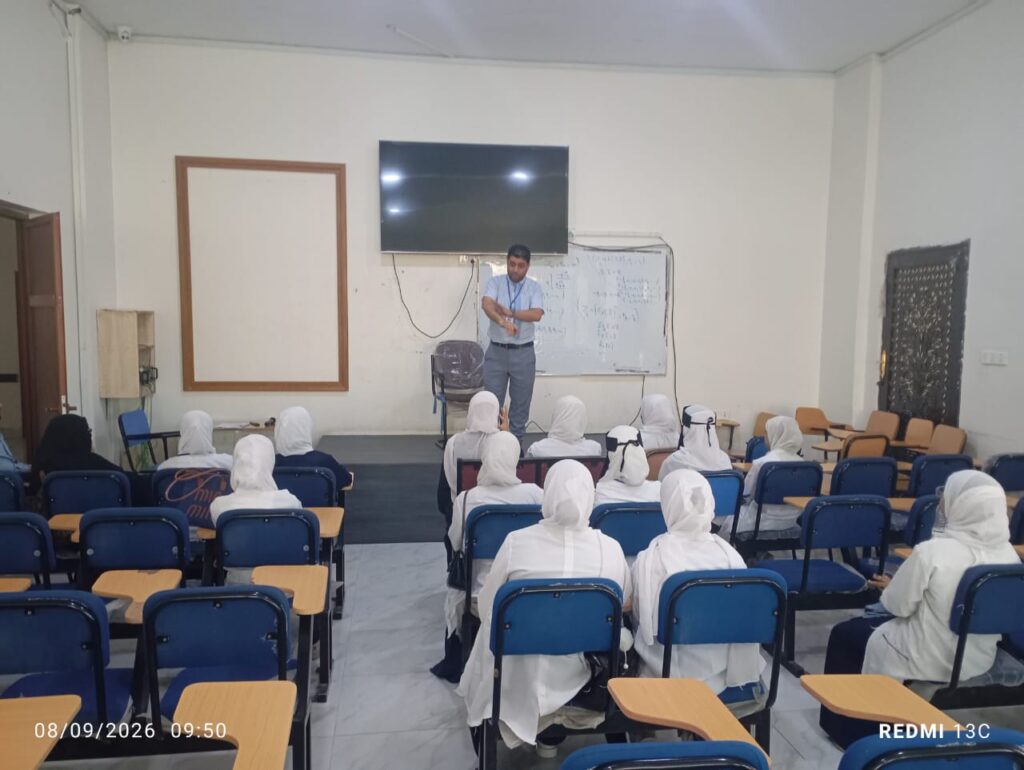 Male instructor explains to a class of female students in white uniforms in a classroom with a mounted TV screen and whiteboard at the front.