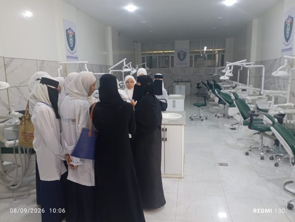 Group of women in white coats and black abayas gathered and talking in a bright dental clinic with green dental chairs along the right.