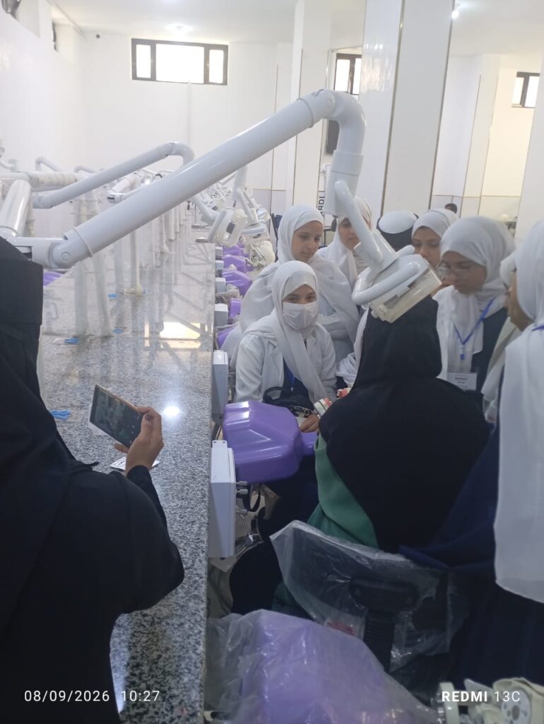 Group of female students wearing white headscarves watch a dental training session around a patient chair with robotic arms in a bright clinic.