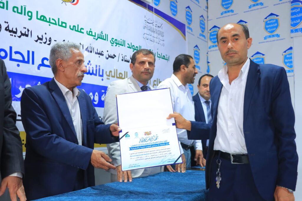 Two men in suits exchange a certificate at a formal ceremony, with a blue banner and logos in Arabic behind them.
