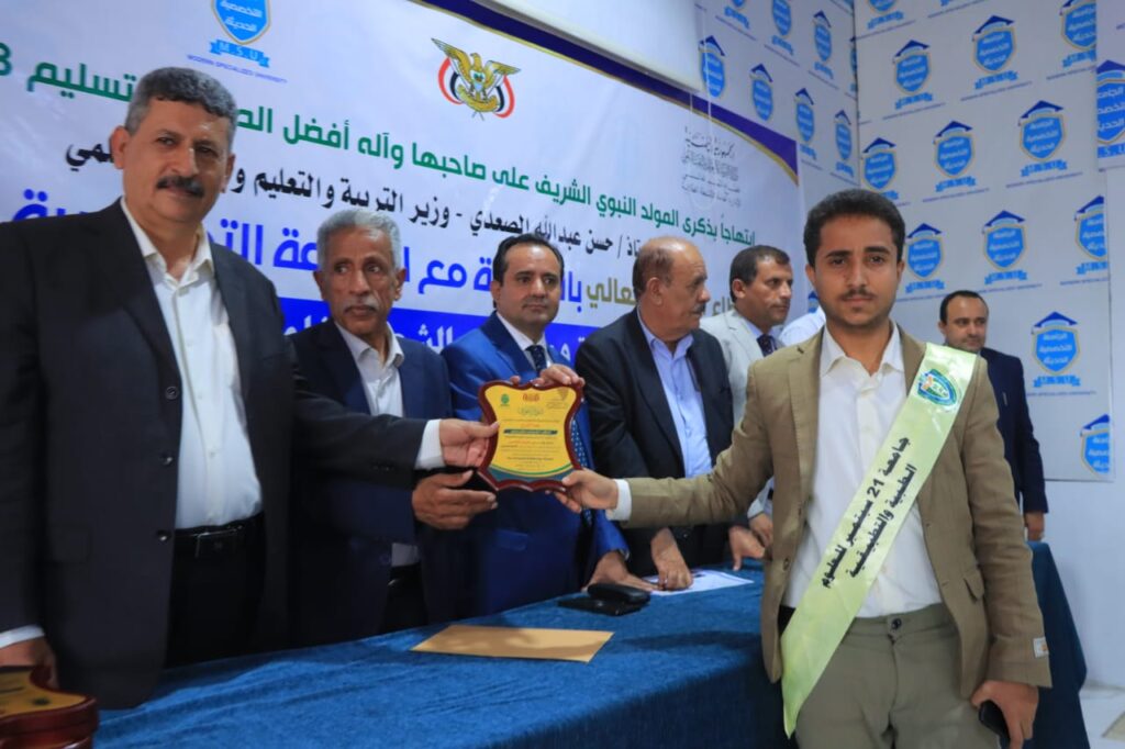 Group of formally dressed men at a ceremony presenting a plaque to a young man wearing a light-colored sash.