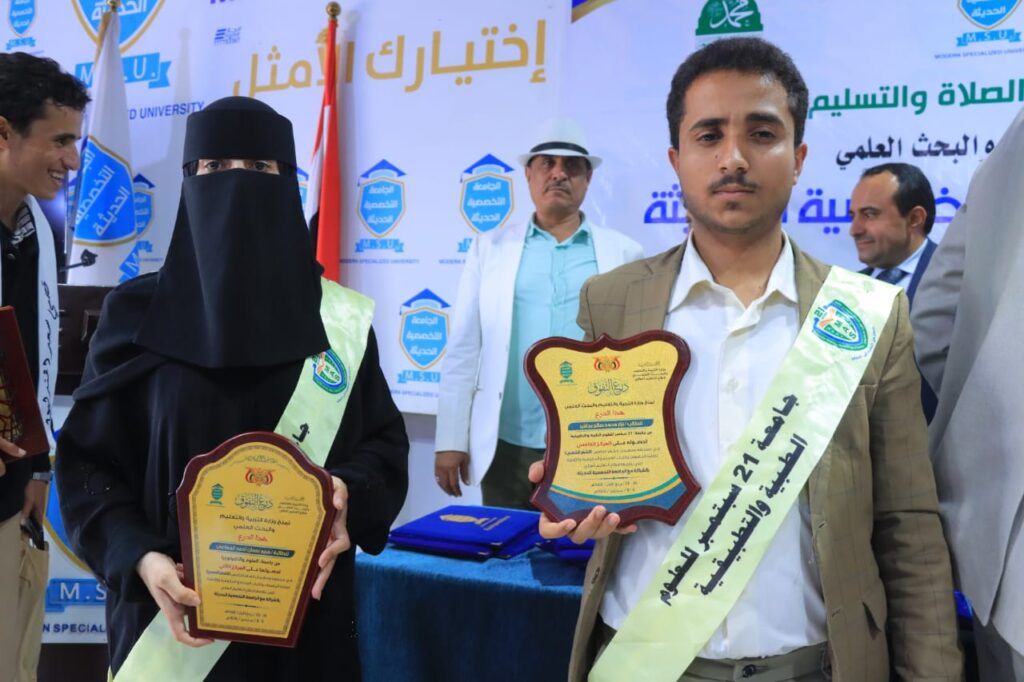 A man and a woman wearing light green sashes hold shield-shaped plaques at a university ceremony, with banners in Arabic in the background.