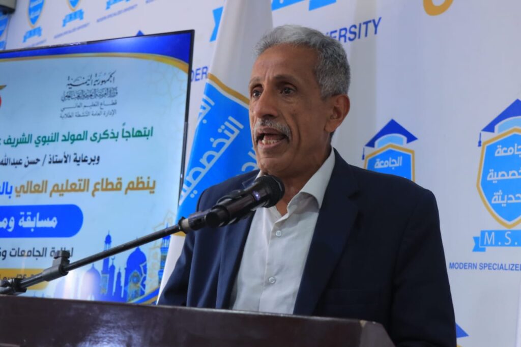 A middle-aged man in a navy suit speaks into a microphone at a podium during a formal event, with a blue and white branded backdrop behind him.
