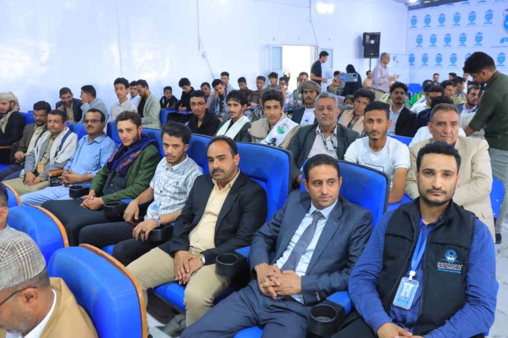 Audience seated in blue theater-style chairs at an indoor event, some attendees wearing badges and lanyards.