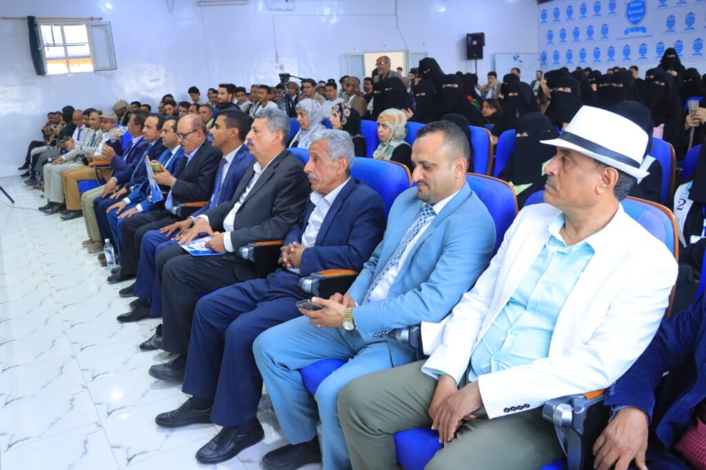 Audience seated in a hall during a formal event; front-row men in suits, background attendees in black abayas.