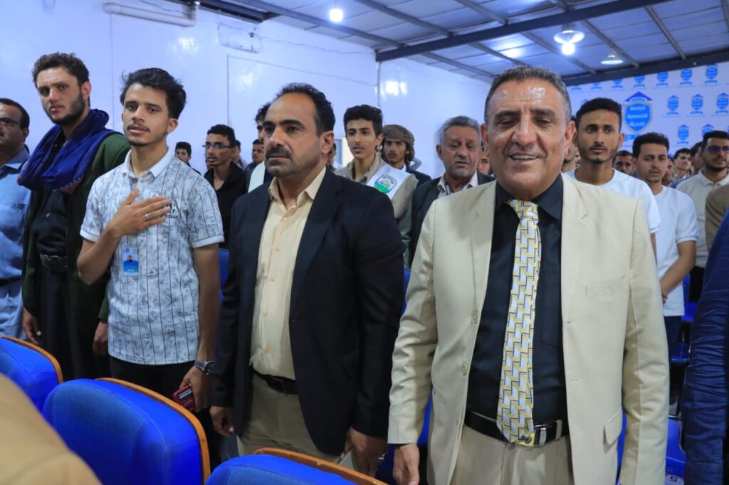 Group of men standing in a conference hall, some with hands over their hearts, attending a formal event; blue chairs and badges visible in the background.