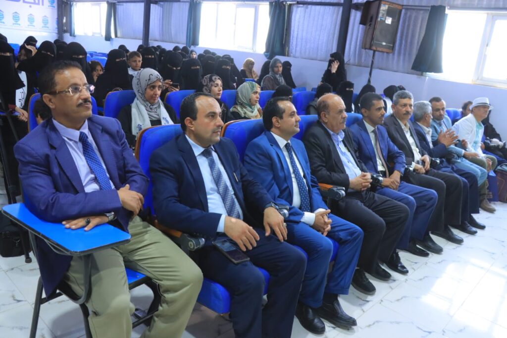 Audience in a conference hall: front row of men in blue suits seated with women in hijabs in the back rows, marble floor, and blue chairs visible.