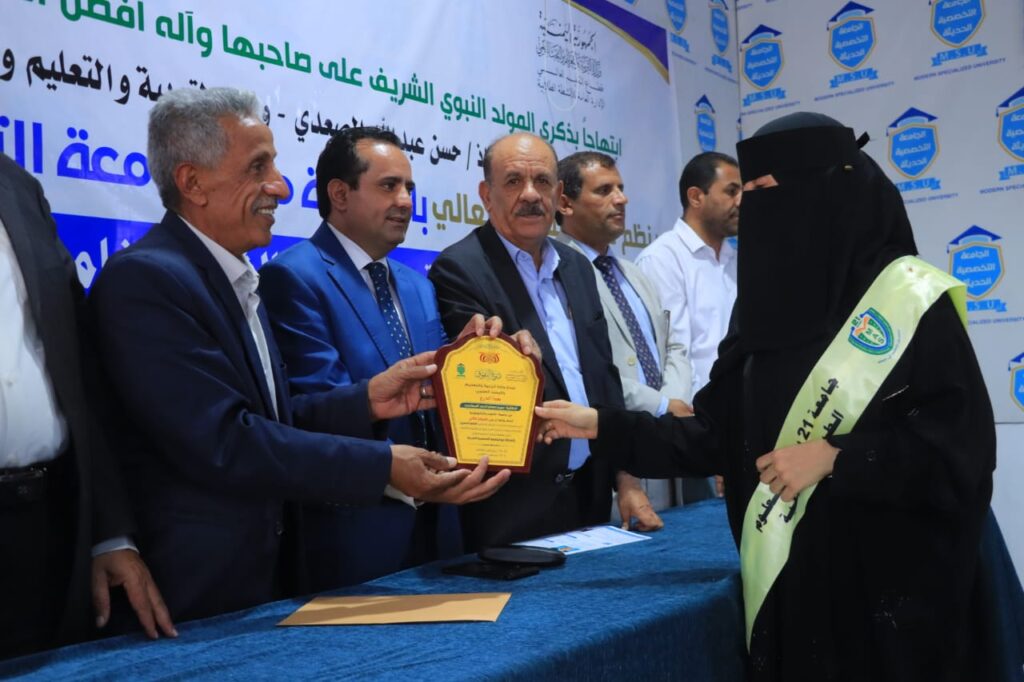 Woman in a black niqab wearing a pale sash receives a yellow plaque from men in suits at an awards ceremony, with a blue banner and Arabic text in the background.