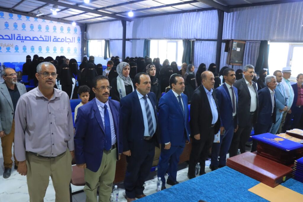 Group of formally dressed men standing at a podium during a ceremony at a university, with women in hijabs seated in the background and a banner on the wall.